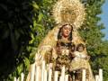 Una estatua de la Virgen María con un niño en sus brazos, rodeada por ramas verdes y una estructura de madera. La escena parece ser en un jardín o parque, con árboles al fondo.