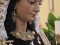 Retrato de una figura vestida con trajes tradicionales, posiblemente religiosos o históricos. La imagen muestra detalles de joyería y adornos, incluyendo una corona y un ancla. La escena parece ser interior, con elementos decorativos como plantas en el fondo.