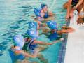 Niñas en traje de baño y gorros de natación azul, nadando en una piscina. Una niña está sentada en la orilla, con el agua hasta sus caderas. La piscina tiene líneas de división y un área de natación con una línea roja.