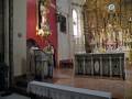 En una iglesia, un sacerdote se prepara para oficiar la misa. En el altar, flores y velas adornan la mesa principal. A la izquierda, un coro se prepara para cantar. La pared roja y dorada contrasta con la arquitectura clásica del templo.