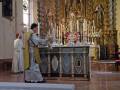 Un sacerdote vestido con un hábito y un manto amarillo se encuentra frente a una mesa altar en una iglesia. El altar está decorado con velas y adornos religiosos, mientras que el sacerdote realiza una acción de oración o bendición. La imagen muestra la belleza y solemnidad típica de una iglesia católica, con detalles artísticos y un ambiente reverente.