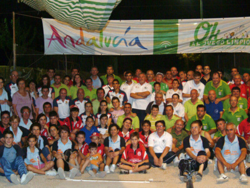 Grupo de personas en una reunión nocturna bajo un toldo, con una bandera de Andalucía y otra de un club deportivo en el fondo.