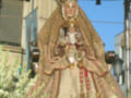 Estatua de la Virgen María vestida con una capa dorada y una corona, en un entorno urbano con edificios y árboles al fondo.