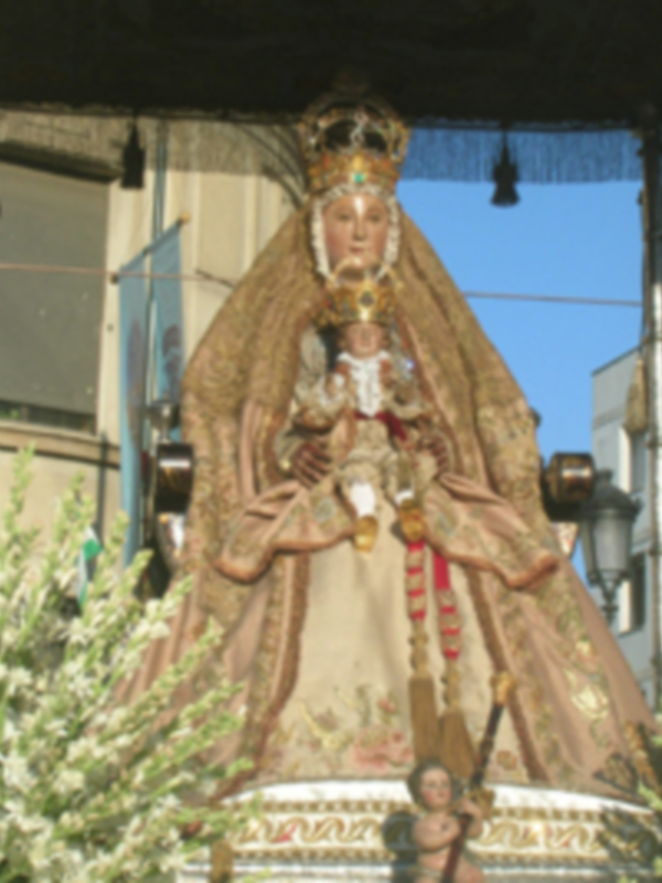 Estatua de la Virgen María vestida con una capa dorada y una corona, en un entorno urbano con edificios y árboles al fondo.