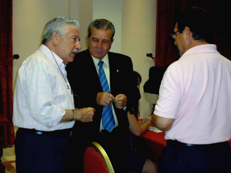 Tres hombres conversan en una reunión formal, uno con camisa a cuadros y otro con traje y corbata.