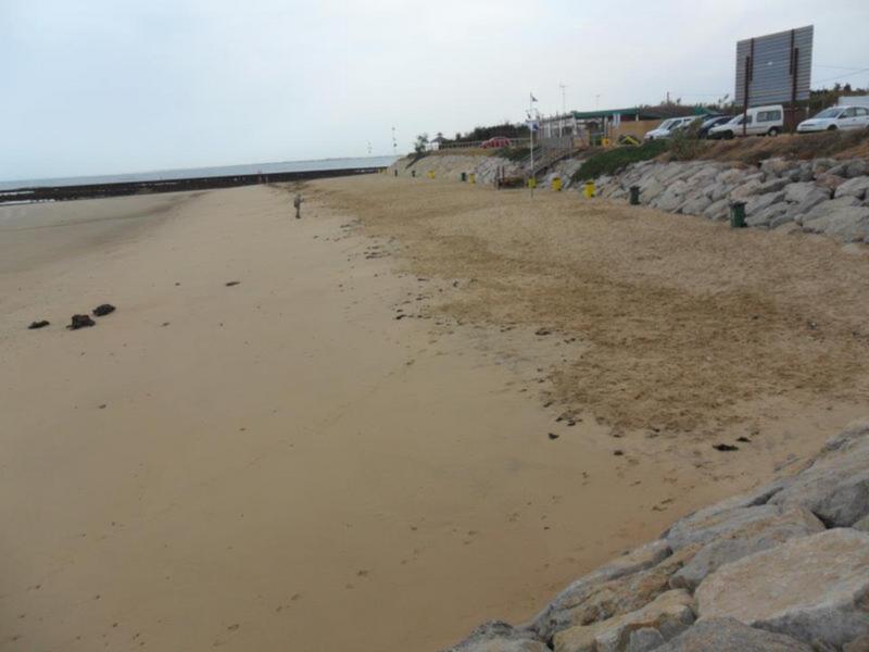 Playa tranquila con arena y rocas en la orilla, con estructuras y vehículos al fondo.