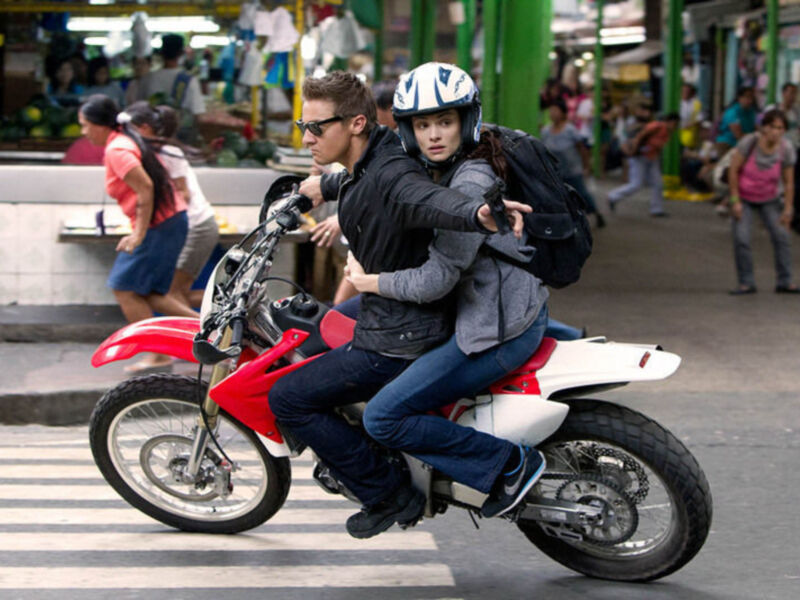 Una pareja monta en una motocicleta roja a través de una calle concurrida, con cascos y ropa casual.