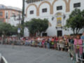 Una multitud de personas se encuentra en una plaza pública, frente a un edificio blanco con detalles arquitectónicos. La gente está sentada y de pie, observando algo fuera del alcance de la imagen. El ambiente es tranquilo y parece ser un evento o reunión importante en el lugar.