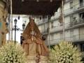 Procesión de la Virgen con carroza ornada y flores blancas en la ciudad.