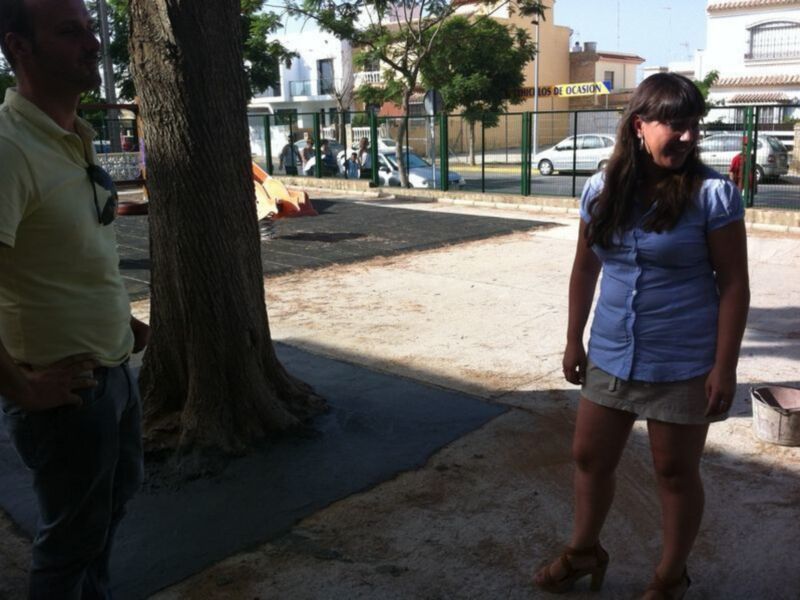 Una joven con un vestido azul posa junto a un hombre en una calle con árboles y edificios en el fondo.