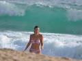 Una mujer en bikini rosa camina por la orilla de una playa con ondas azules y blancas detrás.