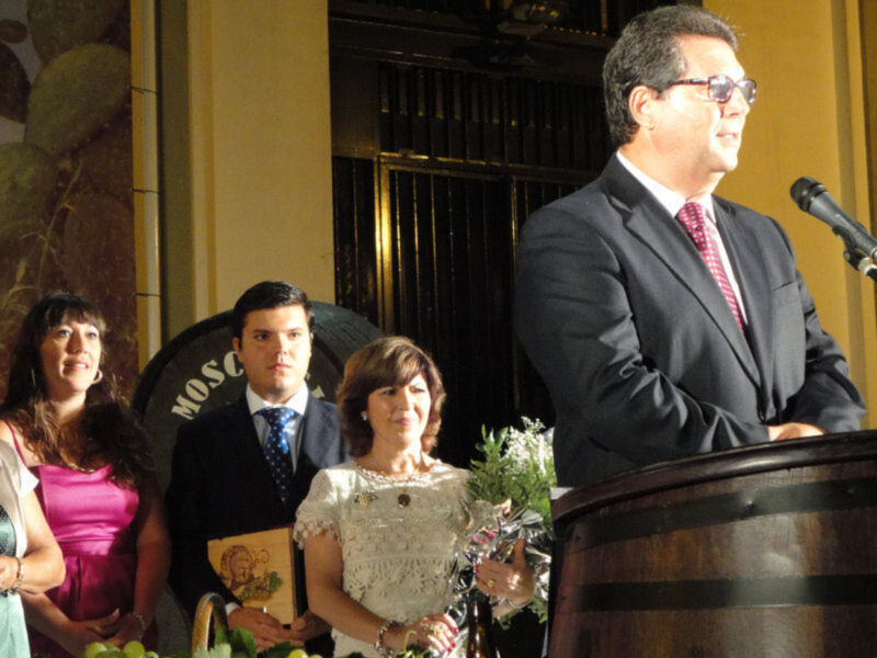 Persona hablando en un evento formal, con un grupo de asistentes detrás en un ambiente con barril de vino y decoración de uvas.