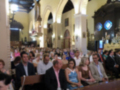 Una multitud en una iglesia, con arcos y vitrales. La imagen muestra a personas sentadas en filas, algunas sonrientes, otros con sombrillas. En el fondo, se observa un altar y una cruz grande.
