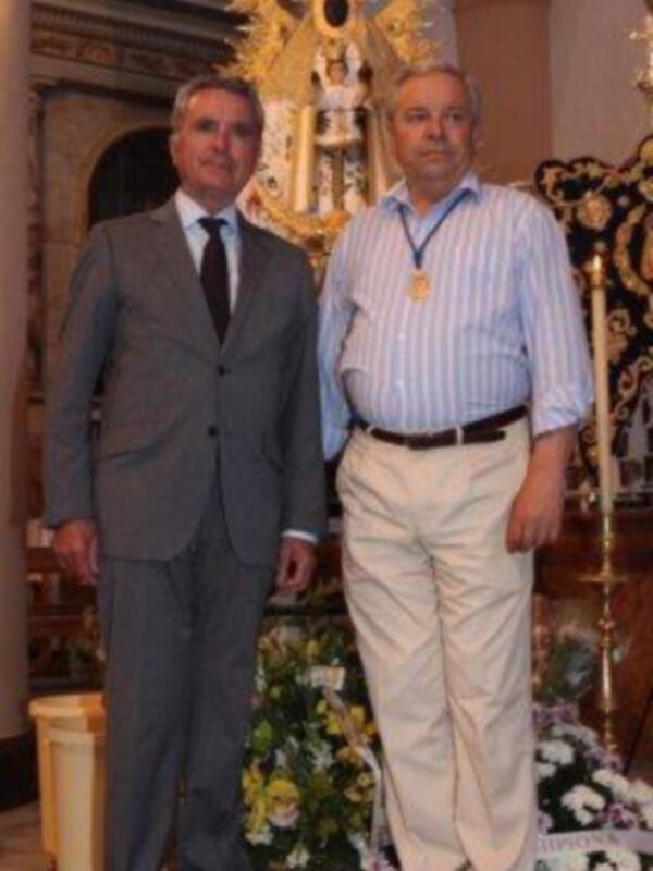 Dos hombres posan juntos en una iglesia, rodeados de decoración festiva y una imagen religiosa en el fondo.