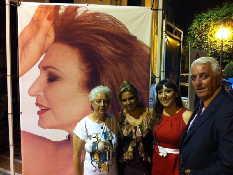 Tres personas posando frente a una gran imagen de una mujer con un fondo de luz amarilla y un árbol en la parte superior derecha.