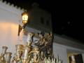 Una procesión nocturna con una estatua de la Virgen María en un carro adornado con candelabros dorados y flores. La escena se ilumina por una lámpara de pared, destacando el ambiente festivo y religioso.