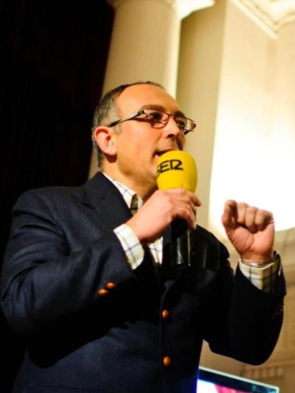 Hombre hablando en un evento formal con un micrófono amarillo.