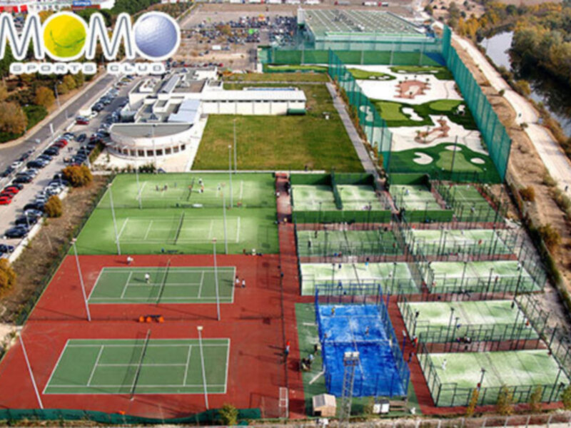 Vista aérea de un club deportivo con pistas de tenis y pádel, rodeado de árboles y edificios.