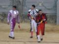 Tres toreros caminan en el ruedo de una plaza de toros. El primero lleva traje rojo y blanco, el segundo un traje negro con detalles blancos y el tercero un traje rojo y negro. Todos llevan guantes amarillos y botas negras.