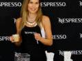 Una mujer con un vestido negro y una camiseta de Nespresso sostiene una taza de café. En la mesa, hay un vaso con un pañuelo y una taza de café. El fondo es negro con el logo de Nespresso repetido.