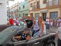 Una multitud de personas celebra en una calle con edificios coloridos. Un grupo saluda desde un auto convertible negro, mientras otros observan la celebración. La imagen captura una festividad en un entorno urbano con edificios de colores y gente vestida casualmente.