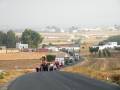 Una caravana de camiones y una carreta con gente en la orilla de una carretera. En el fondo, campos y edificios alineados en la distancia.