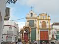 Una procesión religiosa en una calle de la ciudad, con gente observando y participando. En el fondo, una iglesia con un crucifijo en la torre y una imagen de la Virgen. El cielo está nublado, lo que da un tono tranquilo a la escena.