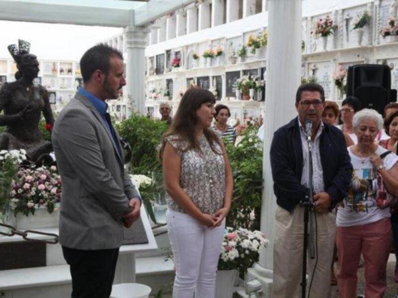 Una ceremonia fúnebre en un cementerio, con una estatua en el fondo y flores en los ataúdes.