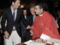 Un hombre en traje negro habla con un sacerdote vestido de rojo, mientras otros hombres observan. En la mesa hay un libro abierto y una pluma.