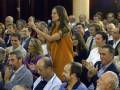 Una mujer en un vestido naranja se levanta con un micrófono, mientras un grupo de personas la aplauden en una sala de conferencias.