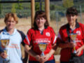 Tres mujeres posan con trofeos en un campo de juego, celebrando su victoria.