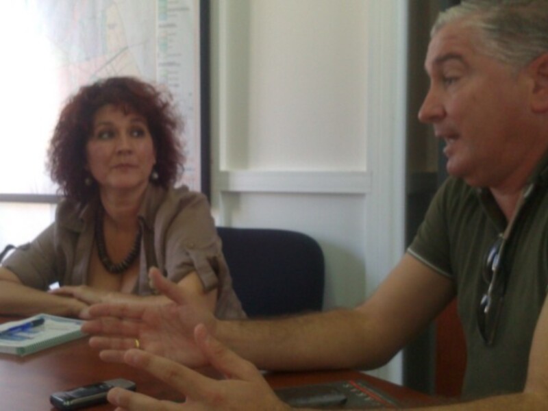 Dos personas en una reunión, discutiendo en un entorno profesional con mapas en el fondo.