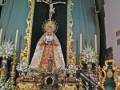 Un altar religioso con una imagen de la Virgen María en el centro, rodeada por flores y adornos dorados. En la parte superior del altar hay una cruz con un Cristo crucificado, y alrededor se pueden ver otros elementos decorativos que incluyen velas y adornos de madera. El fondo es una pared verde con detalles dorados que añaden un toque de elegancia y solemnidad al ambiente.