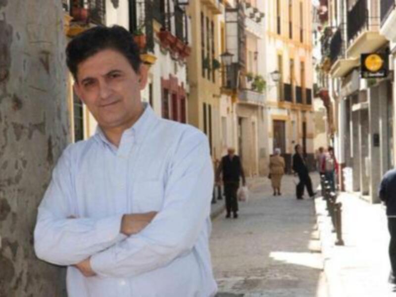 Hombre con camisa blanca y brazos cruzados, posando en una calle adoquinada con edificios antiguos y gente al fondo.