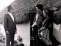 Dos hombres y una mujer conversan en un entorno rural con una casa de techo de paja. La escena es clásica del cine de los años 70, con un estilo cinematográfico que evoca la época.