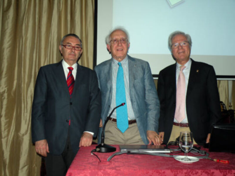 Tres hombres en trajes posan para una foto en un evento formal, con un fondo de cortinas y un proyector en la pared.