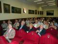 Asamblea en sala de conferencias con asistentes sentados en butacas rojas, retratos en la pared y luces de techo.