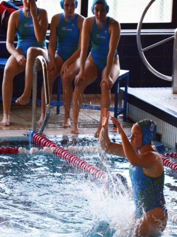 Equipo de natación femenina en acción en una piscina indoor, con una mujer saltando en el agua y otras dos sentadas en el borde del estanque.