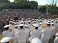 Un grupo de marineros en uniforme blanco toca instrumentos musicales frente a una multitud en un estadio. El ambiente es festivo y el público está atento al desfile musical.