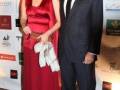Una pareja posa en un evento, la mujer viste una elegante falda roja con un vestido de satén y el hombre lleva traje negro. El fondo muestra logotipos de empresas.