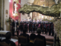 Una procesión religiosa en una iglesia, con un coro de músicos vestidos con trajes tradicionales y un altar dorado en el fondo.