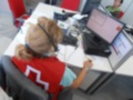 Una persona con cabello rubio y una gorra de béisbol roja está sentada frente a un escritorio con varios dispositivos electrónicos, incluyendo una computadora y un monitor. La persona parece estar trabajando o estudiando en el lugar de trabajo.