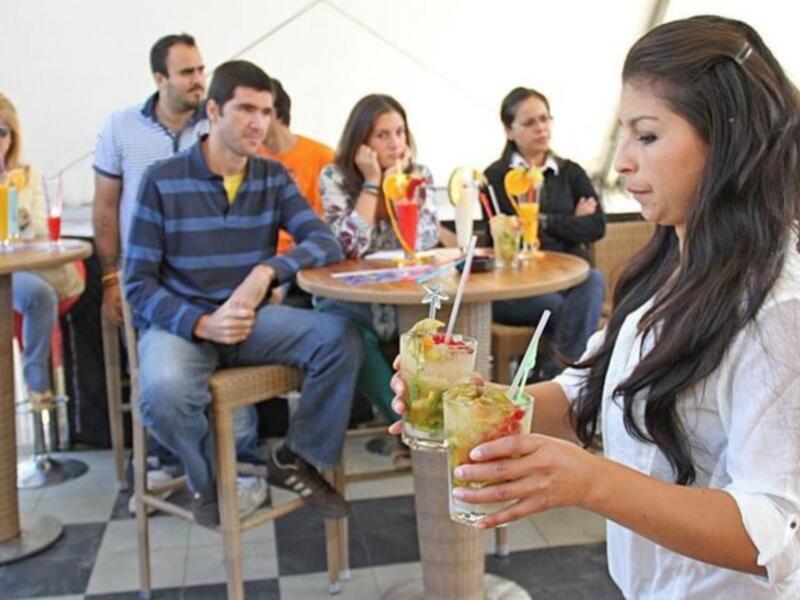 Una camarera sirve cócteles en un ambiente relajado de café al aire libre, mientras clientes disfrutan de su tiempo.