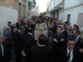 Una multitud de personas en trajes formales se reúne en una calle estrecha, con edificios blancos y una escalera en el fondo.