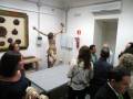 Un grupo de personas observando una exposición de arte en una sala con paredes blancas y un panel con imágenes de figuras antiguas.