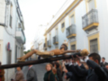 Una imagen de una procesión religiosa en una calle con edificios blancos y amarillos. La gente lleva vestimentas tradicionales, incluyendo capas y sombreros. Una persona sostiene una vara larga con un símbolo religioso en la mano izquierda. La escena transmite una sensación de solemnidad y tradición cultural.