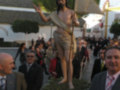 Una escultura de Jesús en la cruz, rodeada por una multitud de personas con trajes formales. El cielo es claro y el ambiente parece ser un evento religioso o festivo en un entorno urbano.