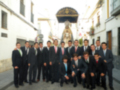 Un grupo de hombres en trajes posando frente a una estatua de la Virgen María en una calle de la ciudad.
