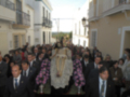 Una procesión religiosa con personas vestidas de negro, llevando una imagen de la Virgen María en una cruz. La escena se desarrolla frente a edificios blancos y amarillos, con un cielo claro.