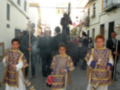 Tres niños vestidos con trajes tradicionales marchan en una procesión, seguidos por adultos. La imagen muestra un ambiente festivo con decoraciones y gente en la calle.
